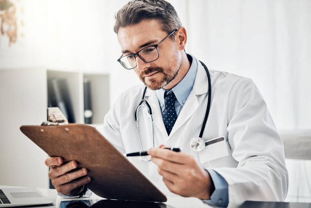 Patienten haben für ihn oberste Priorität. ein fokussierter Arzt betrachtet Papierkram auf einem Klemmbrett, während er an seinem Schreibtisch sitzt - Foto, Bild