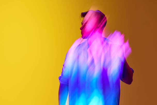 Young stylish man standing against gradient yellow background with mixed neon multicolored lights effect. Futurism. Concept of modern photography, art, cyberpunk, techno, creativity, beauty - 写真・画像