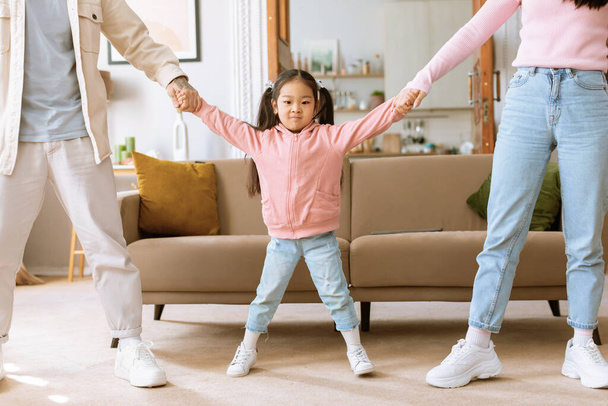 Családi összetartás. Vicces kis japán kölyök lány fogja a kezét apuval és anyuval, nézi a kamera pózol a szülők állnak a modern nappaliban otthon. Vágott lövés - Fotó, kép