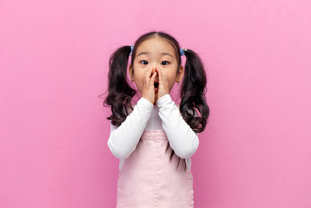 little shocked Asian girl in pink sundress with long hair is surprised and covers her mouth with her hands on pink isolated background, amazed Korean preschool child screams in shock - Zdjęcie, obraz