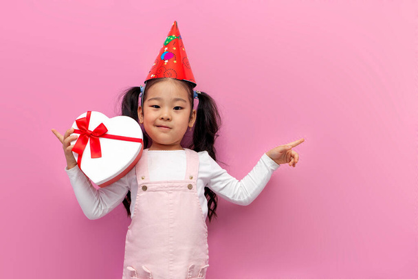 little asian girl celebrates birthday in party cap and holds gift heart on pink isolated background and points to the side with her hand, surprised korean child in festive outfit advertises copy space - Foto, Bild