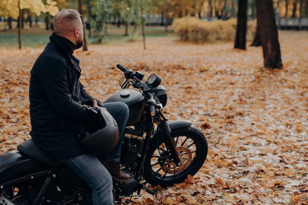 Widok z tyłu brodatego motocyklisty patrzy troskliwie gdzieś w oddali, pozuje na czarny motocykl, trzyma kask ochronny, spędza czas wolny w jesiennym parku, kopiuje miejsce na reklamę - Zdjęcie, obraz