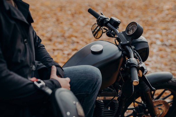 Cropped shot of faceless biker poses on fast motorbike, stops in park, ground covered with fallen leaves, enjoys travel and journey. People, lifestyle, transport concept. - Photo, Image