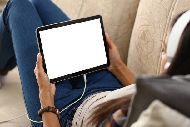Mockup beeld van een digitale tablet met zwart scherm in handen van vrouw in hoofdtelefoon. Persoon die thuis op de bank rust met tablet over schouderzicht - Foto, afbeelding