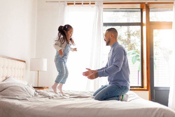 kis ázsiai lány ugrás az ágyon otthon apával, koreai férfi játszik a lányával, fiatal boldog család, apa szerelem koncepció - Fotó, kép