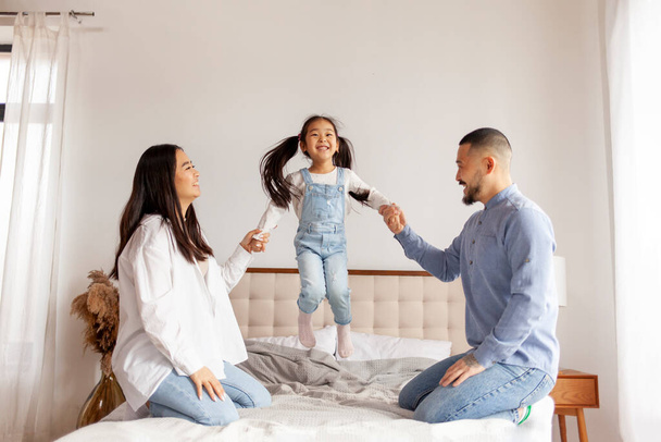 Ευτυχισμένη ασιατική οικογένεια με την κόρη παίζουν και χαίρονται στο σπίτι στο κρεβάτι, μικρό κορεάτικο κορίτσι άλμα στο κρεβάτι και κρατώντας τα χέρια μαμά και ο μπαμπάς - Φωτογραφία, εικόνα