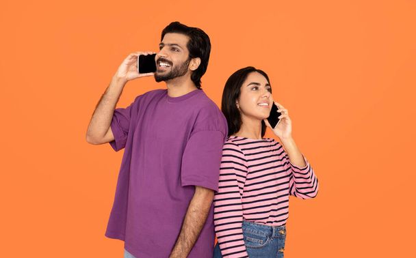 Aplicación móvil. Hermosa alegre feliz amante joven pareja india en trajes casuales de pie espalda con espalda, el uso de teléfonos celulares, tener conversación sobre fondo de estudio naranja, mirando espacio de copia - Foto, Imagen