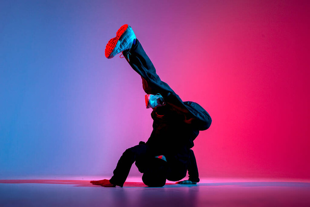 young guy dancer is dancing break in neon lighting, male acrobat is doing trick and dance exercise on red blue background, hip hop performer is moving and standing on his hands - Photo, image
