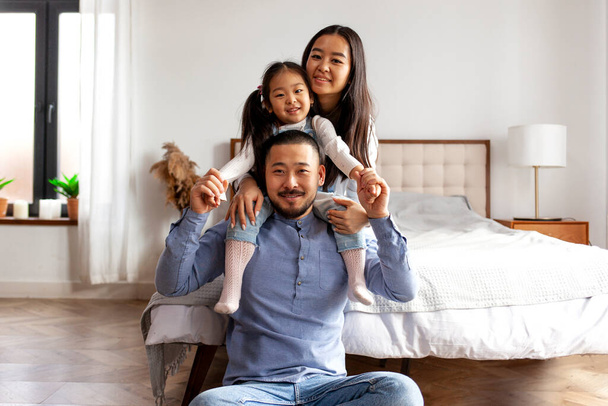 щаслива азіатська сім'я з маленькою донькою сидить вдома біля ліжка і посміхається, корейська дівчина сидить з татом і мамою, батьківська любов і догляд
 - Фото, зображення