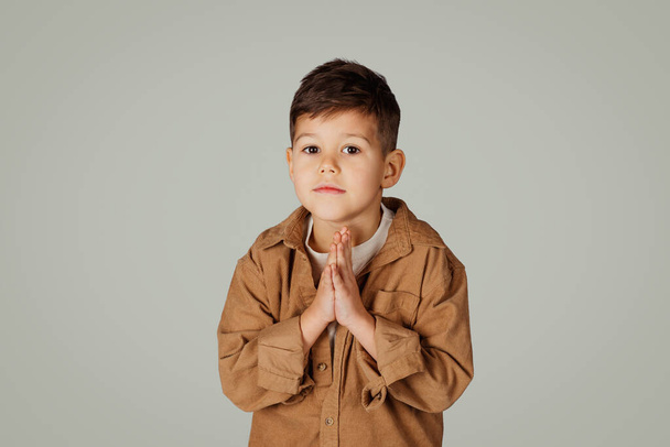 Niño serio de 6 años en casual hace gesto de oración, pide gesto aislado sobre fondo gris, estudio, espacio de copia. Infancia, estudio, emociones de los alumnos, anuncio y oferta - Foto, Imagen