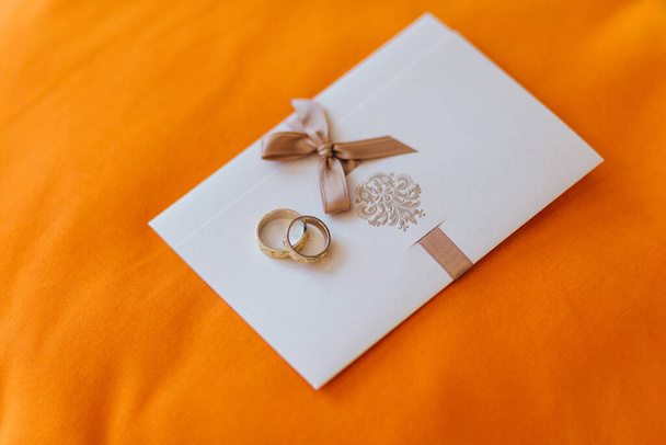 Hochzeitskonzept. Goldene Eheringe liegen auf einer weißen Einladungskarte vor orangefarbenem Hintergrund. Ehe und Heiratsantrag. Einladung zu besonderen Anlässen - Foto, Bild