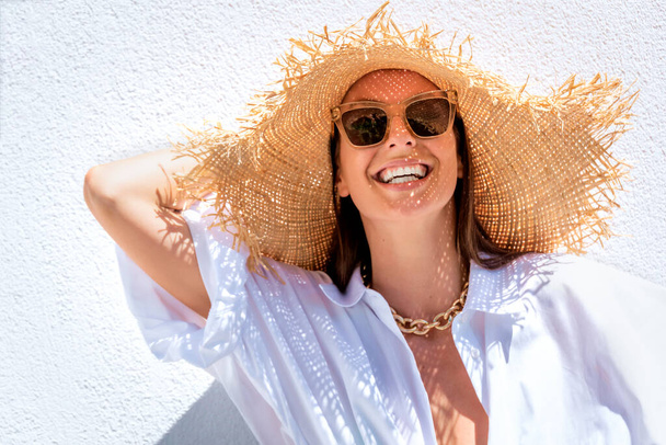 Close-up of an attractive woman wearing sunhat and sunglasses while standing at white wall. Happy woman enjoying sunshine. - 写真・画像