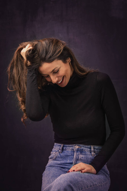 Attractive brunette haired woman wearing black sweater and jeans while standing at isolated dark background.  - Фото, изображение