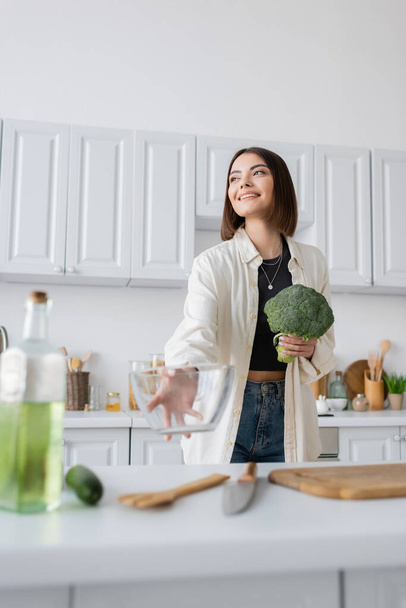 Positive woman holding broccoli and blurred bowl in kitchen  - Valokuva, kuva