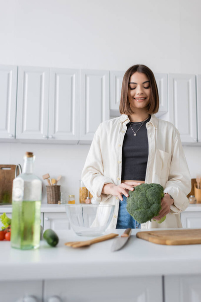 Overjoyed woman holding broccoli near cutting board in kitchen  - Fotografie, Obrázek