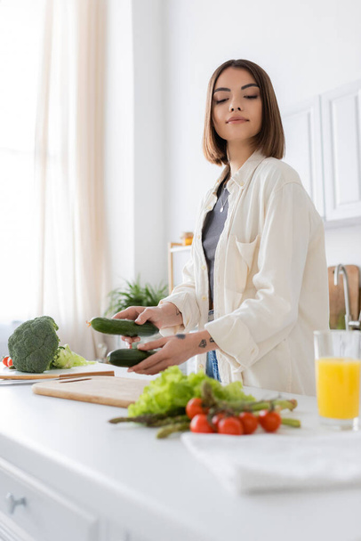 Brunette woman holding cucumbers near blurred vegetables and orange juice in kitchen  - Foto, afbeelding