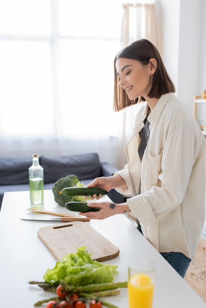 Uśmiechnięta kobieta trzyma ogórki w pobliżu warzyw i deski do krojenia w kuchni  - Zdjęcie, obraz