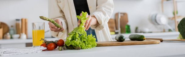 Cropped view of young woman holding asparagus and lettuce in kitchen, banner  - Foto, Bild