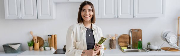Carefree woman looking at camera and holding asparagus in kitchen, banner  - Foto, Bild