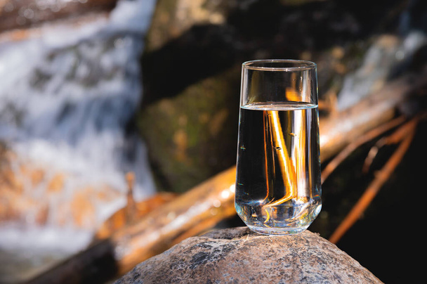 A glass of water at a waterfall. A glass glass with drinking water stands on a stone in front of a waterfall, a blurred background. Pure drinking mineral water. - Фото, зображення