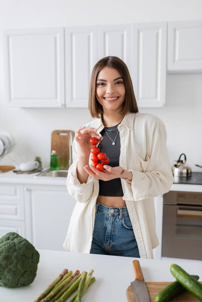 Smiling young woman holding cherry tomatoes near vegetables in kitchen  - Fotoğraf, Görsel