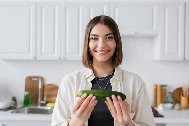Femme brune souriante tenant le concombre et regardant la caméra dans la cuisine  - Photo, image