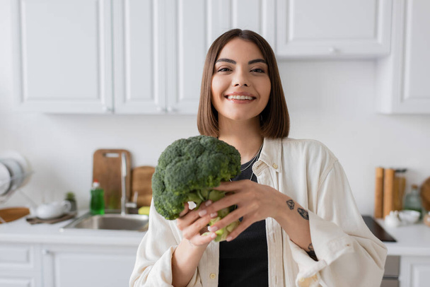Positive young woman looking at camera while holding fresh broccoli in kitchen  - Fotoğraf, Görsel