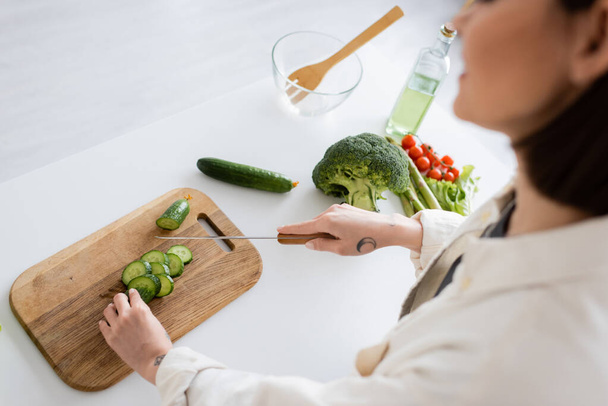 Cropped view of blurred woman cutting cucumber near fresh vegetables in kitchen  - Foto, immagini