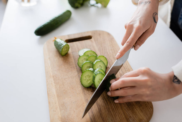 Cropped view of tattooed woman cutting cucumber on board in kitchen  - Photo, Image
