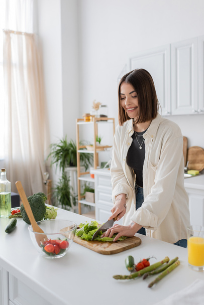 Positive brunette woman cutting lettuce while cooking salad in kitchen  - Fotó, kép