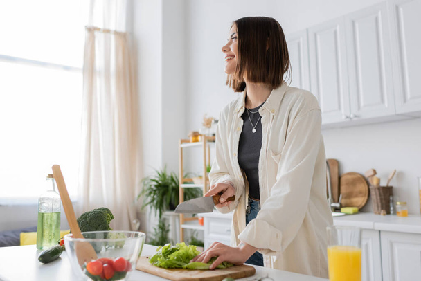 Усміхнена молода жінка розрізає салат біля апельсинового соку та салату на кухні
  - Фото, зображення