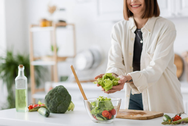 Cropped view of smiling woman putting lettuce in bowl with salad in kitchen  - Fotoğraf, Görsel