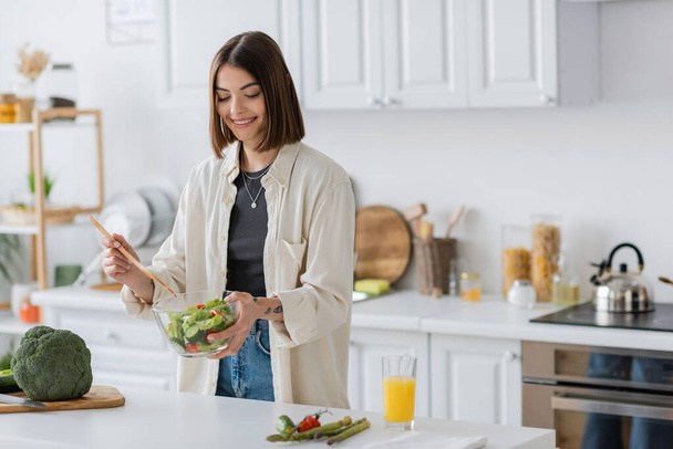 Donna sorridente che tiene ciotola con insalata fresca in cucina a casa  - Foto, immagini