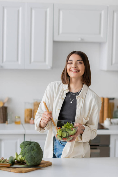 Positive woman mixing fresh and vegetarian salad in kitchen  - Photo, image