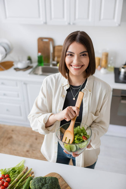 Pozytywna młoda kobieta trzyma świeżą sałatkę i patrzy w kamerę w kuchni  - Zdjęcie, obraz