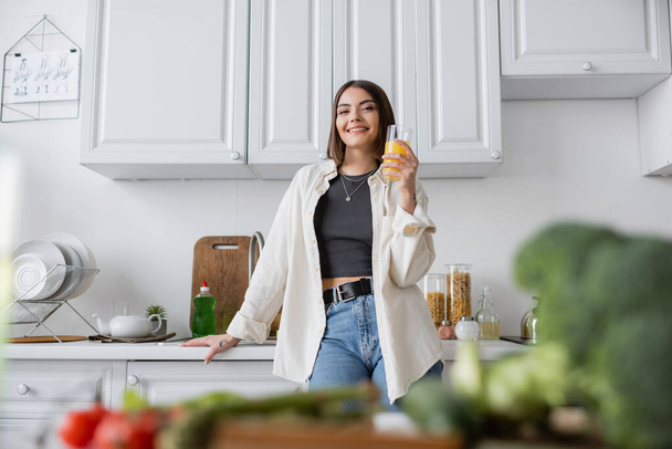 Positive woman holding orange juice near blurred vegetables in kitchen  - Fotó, kép