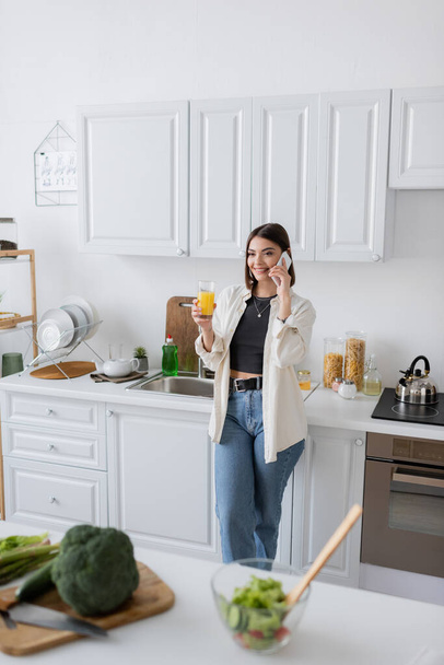 Cheerful young woman talking on cellphone and holding orange juice in kitchen  - Photo, image