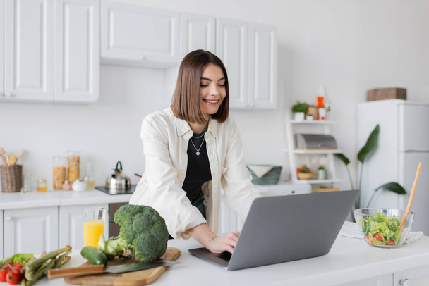 Positive woman using laptop near ripe vegetables and orange juice in kitchen  - 写真・画像