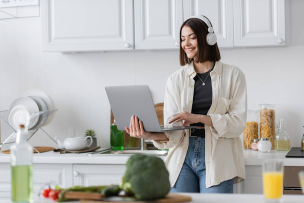 Carefree woman in headphones using laptop near blurred vegetables and olive oil in kitchen  - Foto, Imagem