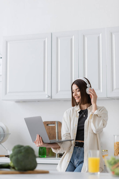 Mulher positiva em fones de ouvido usando laptop perto de brócolis borrado e suco de laranja na cozinha  - Foto, Imagem