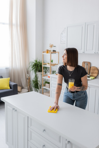 Vue latérale de la femme tatouée souriant et tenant du jus d'orange tout en nettoyant le plan de travail dans la cuisine  - Photo, image