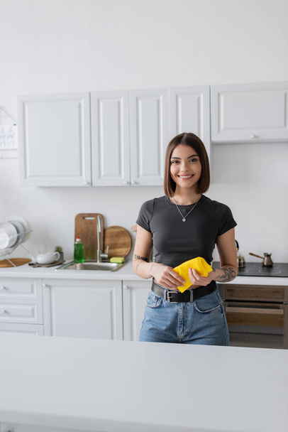 Smiling tattooed woman holding rag and looking at camera in kitchen  - Foto, imagen