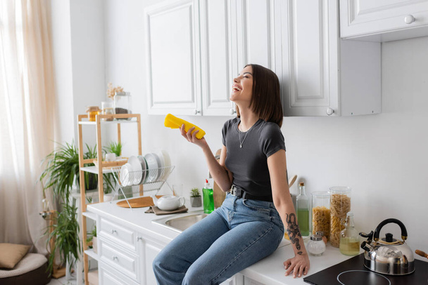 Overjoyed tattooed woman holding rag while sitting on worktop in kitchen  - Photo, image