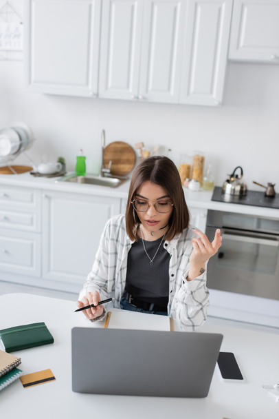 Freelancer in eyeglasses using laptop near notebooks and credit card in kitchen  - Foto, Imagen