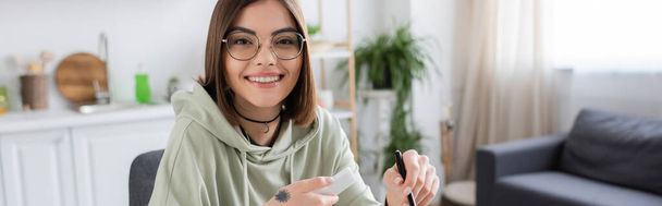 Smiling woman in eyeglasses holding smartphone and pen at home, banner  - Foto, Imagen