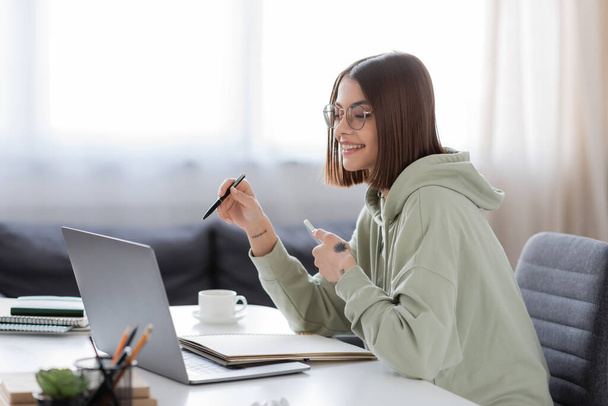 Позитивный фрилансер в очках держит смартфон возле ноутбука и кофе дома  - Фото, изображение
