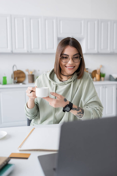 Cheerful freelancer holding coffee cup near blurred laptop and credit card at home  - Valokuva, kuva