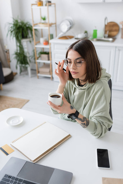 Tattooed woman in eyeglasses holding coffee cup near devices and credit card on table at home  - Foto, Bild