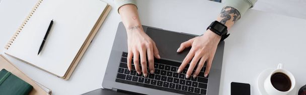 Vista superior do freelancer tatuado usando laptop perto de notebooks e xícara de café na mesa, banner  - Foto, Imagem
