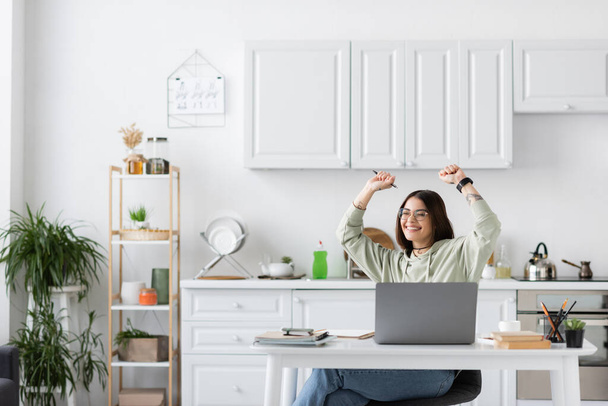Emocionado freelancer mostrando sí gesto cerca de la computadora portátil y portátiles en la mesa en la cocina  - Foto, Imagen